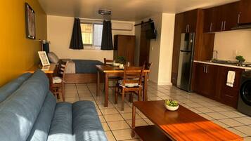 A living room with a sofa, dining table, chairs, and a kitchen area with cabinets and appliances.