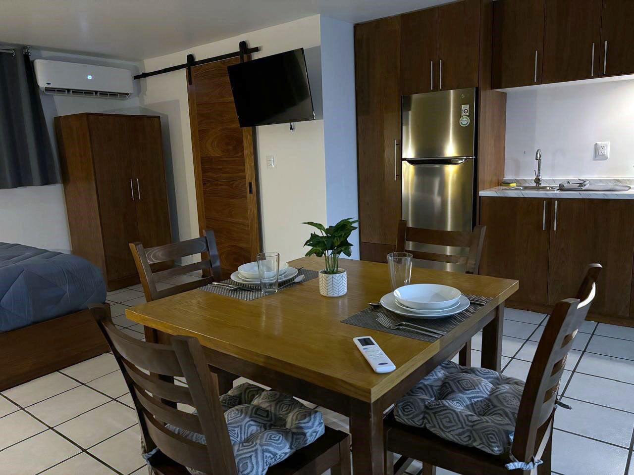 Habitación de hotel pequeña con una mesa de comedor de madera dispuesta para cuatro personas, una cama con cubrecama azul, un televisor montado en la pared y una kitchenette con refrigerador de acero inoxidable.