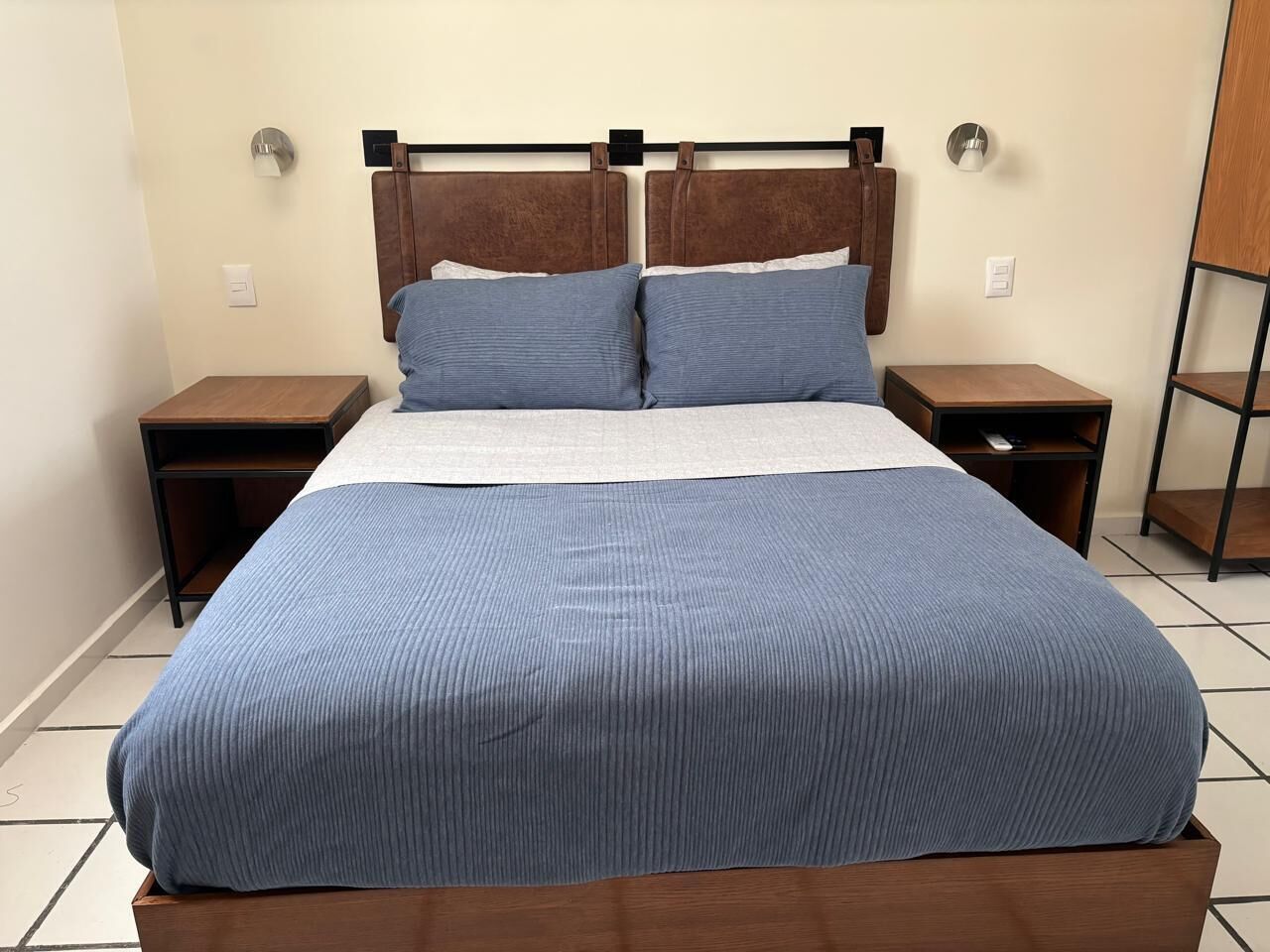Una cama con estructura de madera, dos mesitas de noche y cabecera con luces fijadas a la pared.