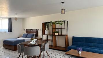 A modern, well-lit room with a bed, a sofa, a dining table with chairs, and a small coffee table. There is a bookshelf with plants and a hanging light fixture.