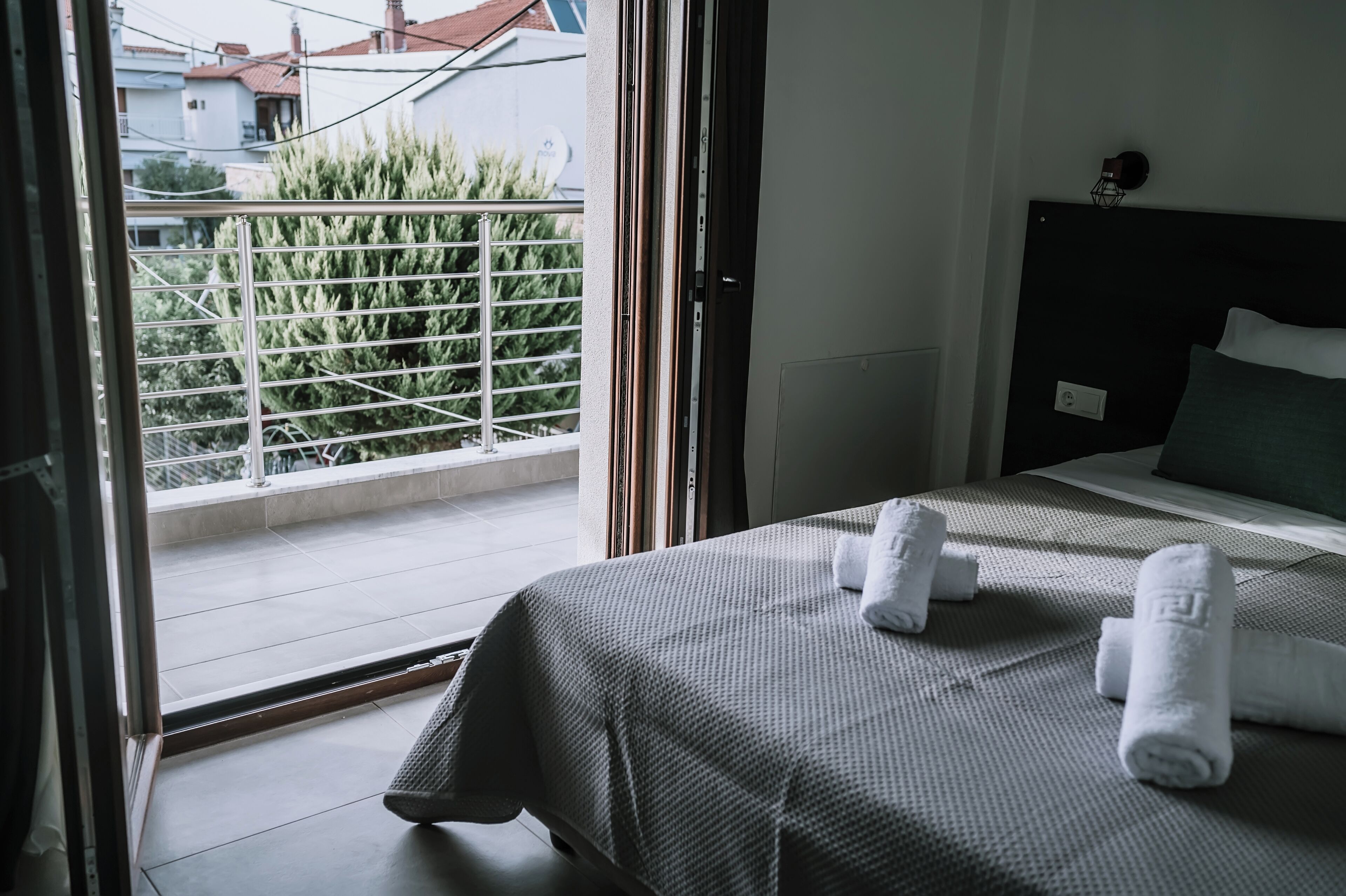 Une chambre d’hôtel avec un lit, deux serviettes enroulées, un balcon doté d’une rambarde en métal et une vue sur la verdure et les bâtiments environnants.
