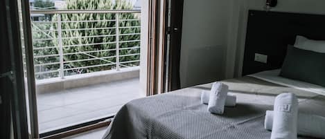 A hotel room with a bed, two rolled towels, a balcony with a metal railing, and a view of greenery and buildings outside.