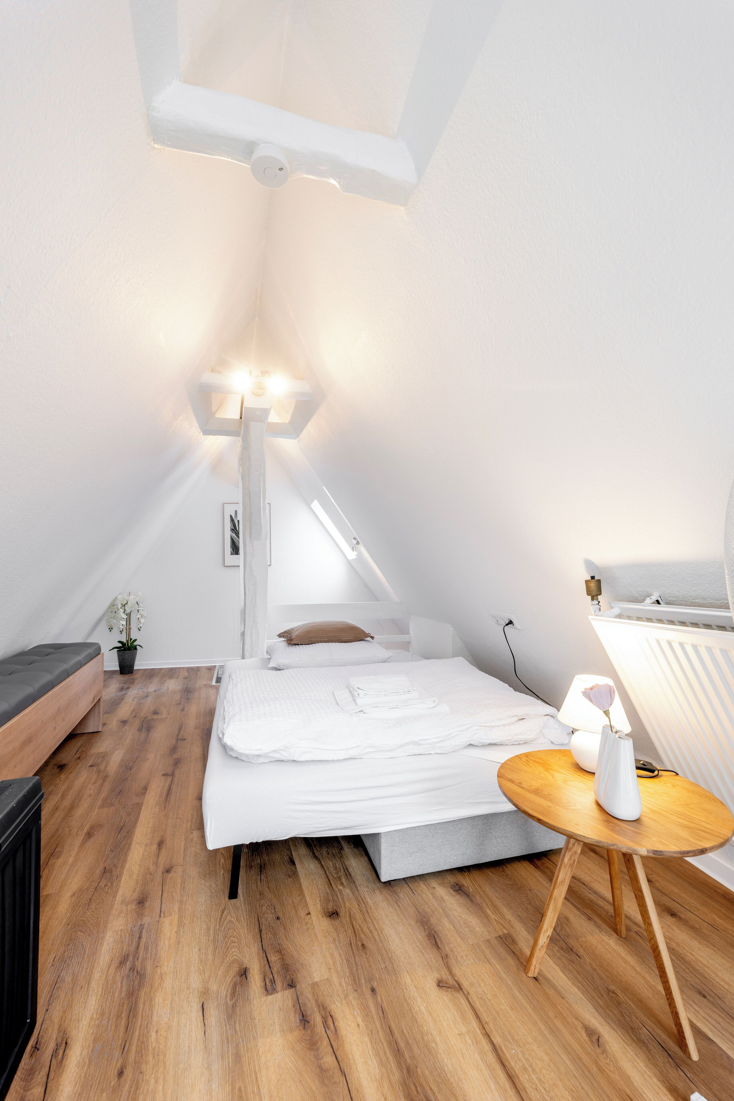 Ein modernes, helles Schlafzimmer im Dachgeschoss mit Holzboden, einem Bett mit weißer Bettwäsche, einem kleinen runden Tisch und einer Nachttischlampe.