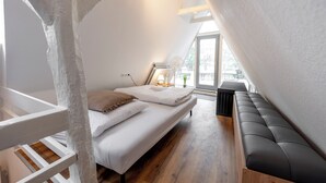 Un dormitorio con cama alta, un banco y ventana que deja entrar luz natural.