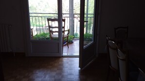 Una habitación con piso de madera, una mesa de comedor con sillas y un vidrio corredizo que da a un balcón con una silla y plantas en macetas.