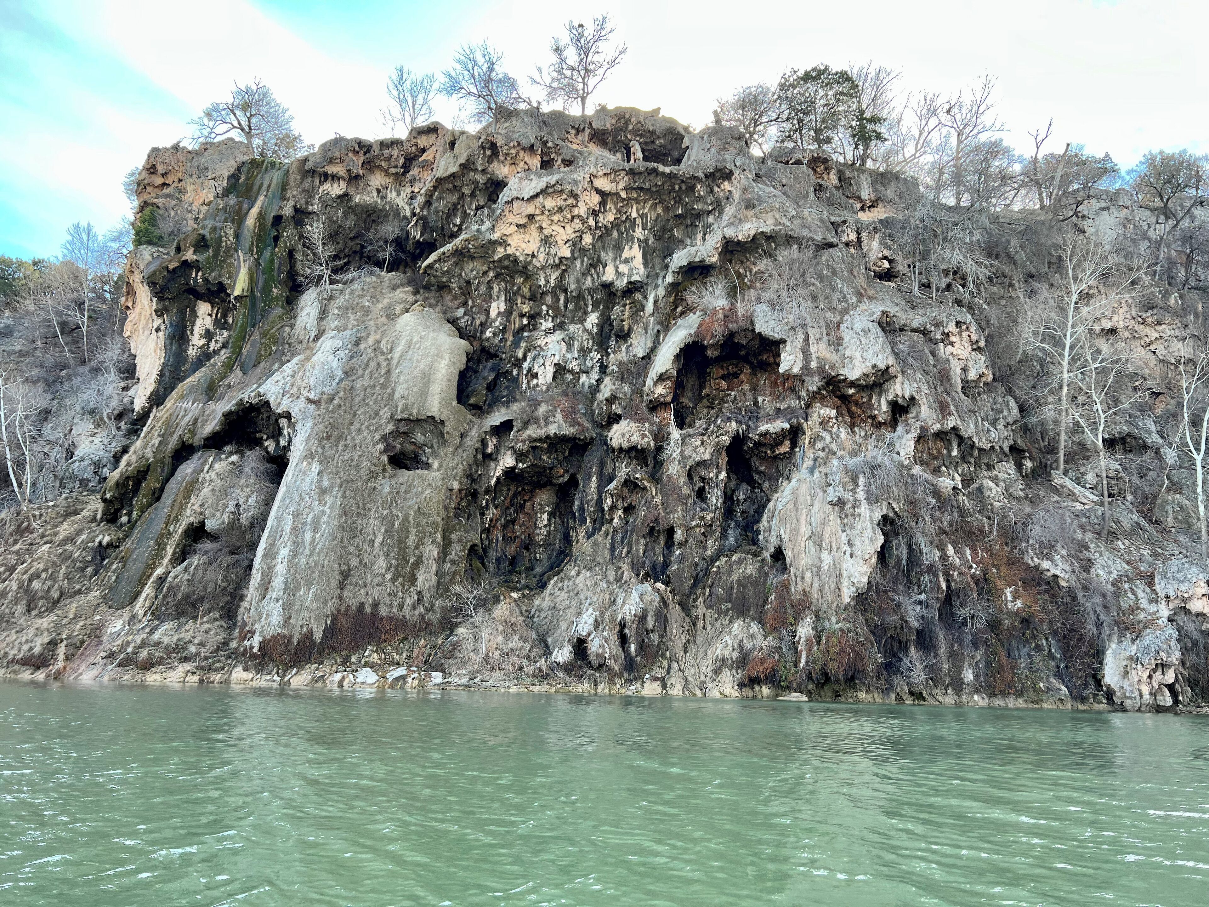 A rocky cliff with multiple caves and a river in the foreground.