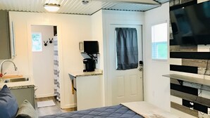 Een compacte slaapkamer met een bed, een klein keukentje, een televisie en een deur naar een andere kamer.
