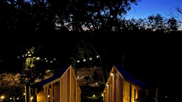 Un puente de madera con barandillas e iluminación, que conduce a un edificio con muro de piedra y un árbol al fondo.