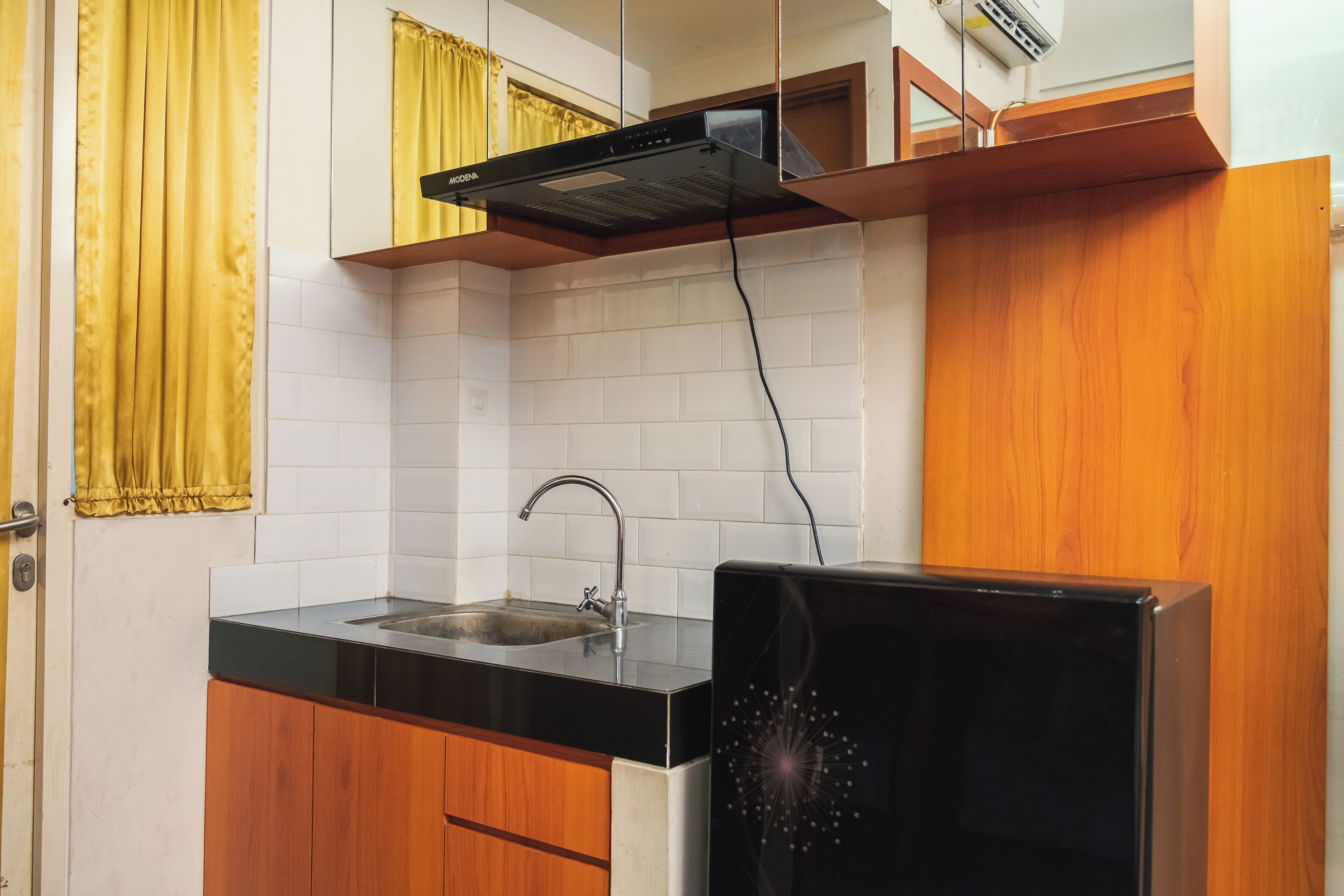 A kitchen with a black countertop, a sink, wooden cabinets, a black range hood, and a yellow curtain.