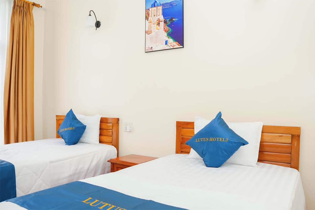 Hotellihuoneessa on kaksi sänkyä, joissa kummassakin on sininen tyyny ja sängynpeite, jossa on hotellin nimi. Sänkyjen välissä on yöpöytä, seinälle kiinnitetty lamppu ja sänkyjen yläpuolella on taulu, jossa on kuvattuna merimaisema.
