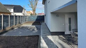 Een smalle binnenplaats met een geplaveid pad, een metalen poort en een wit gebouw met een deur en een klein raam.