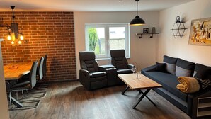 Een moderne woonkamer met een bakstenen muur, een eethoek met een houten tafel, twee leren fauteuils, een zwarte bank en een houten salontafel.