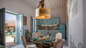 Un coin repas avec une table en verre, des chaises en bois et un élément décoratif central en bois. On aperçoit à l’arrière-plan une cuisine avec des armoires de couleur turquoise et un pare-feu en pierre.