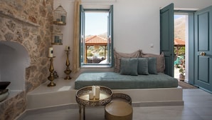 Une pièce chaleureuse avec un mur en pierre, un coin fenêtre agrémenté de coussins, une petite table avec des bougies, et une vue sur une montagne et des bâtiments aux toits rouges.