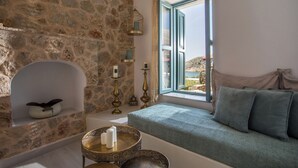 Une pièce aux murs de pierre, avec une fenêtre, un banc rembourré, une petite table avec des bougies et une lampe décorative.