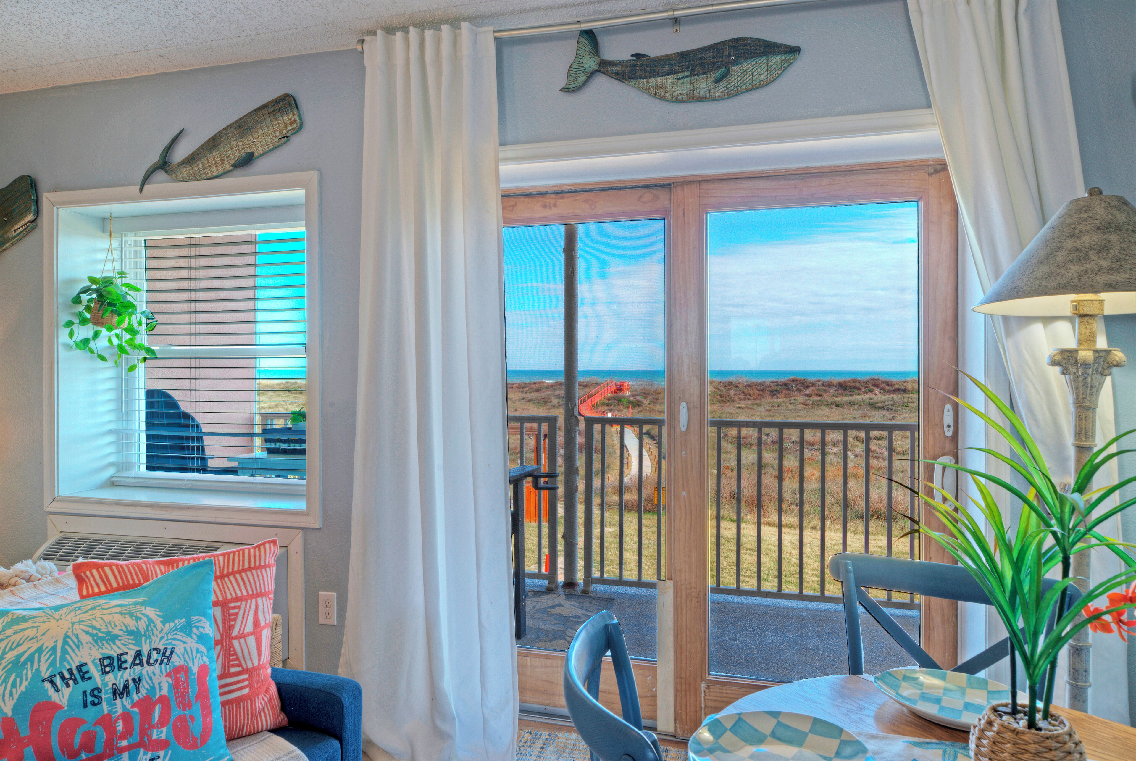 Quarto com vista para a praia, uma cadeira azul, uma mesa com toalha xadrez e um abajur.