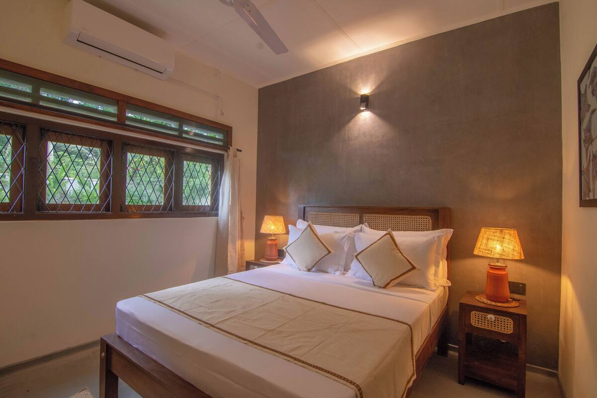 A neatly made bed with a wooden headboard, two bedside lamps, and a window with a view of greenery.