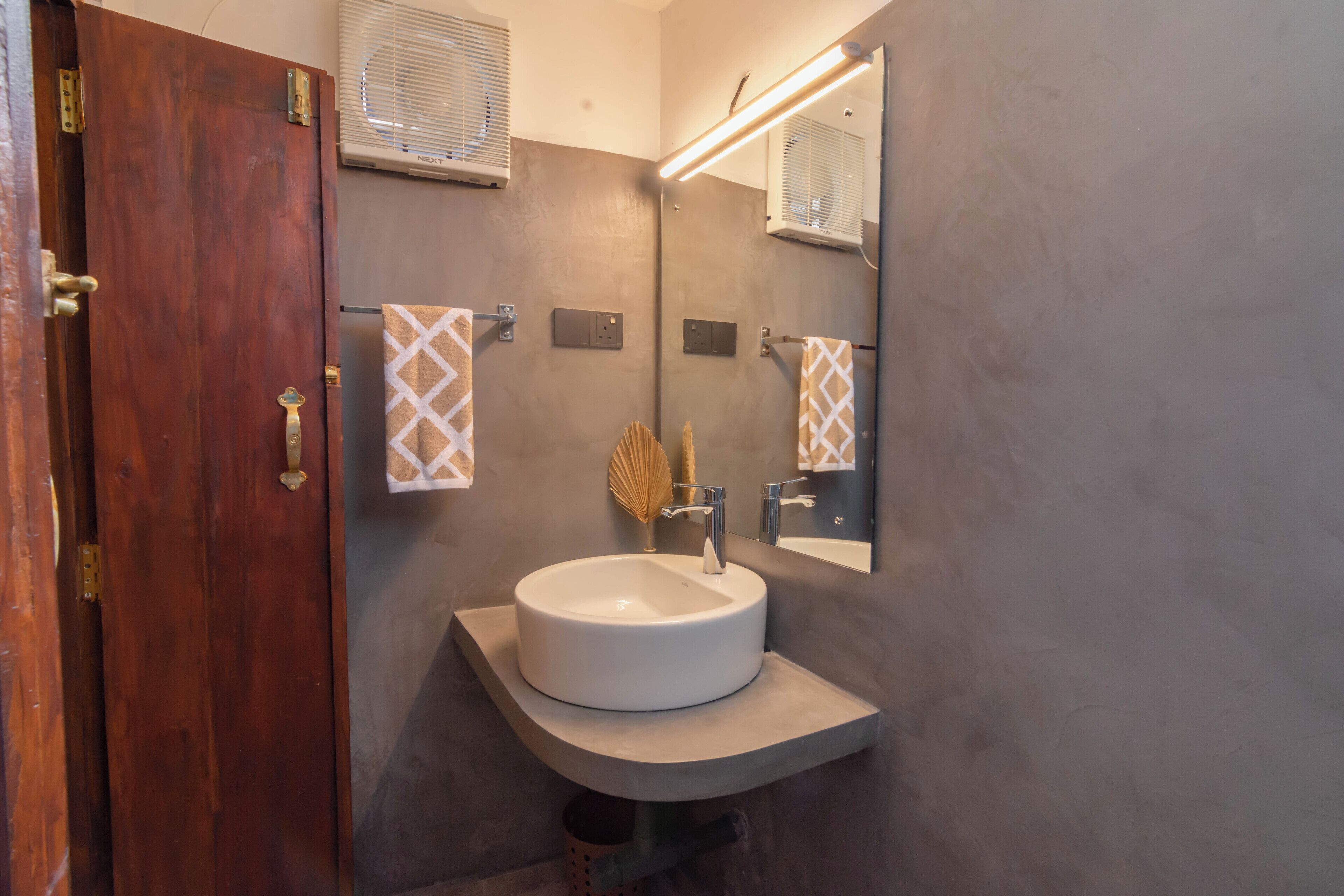 Une salle de bain avec une porte en bois, un lavabo blanc, un porte-serviettes avec des serviettes à motifs, un miroir et un climatiseur fixé au mur.
