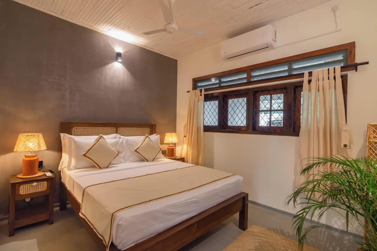 A neatly made bed with white linens and a wooden headboard, two bedside lamps, a window with curtains, and a ceiling fan.