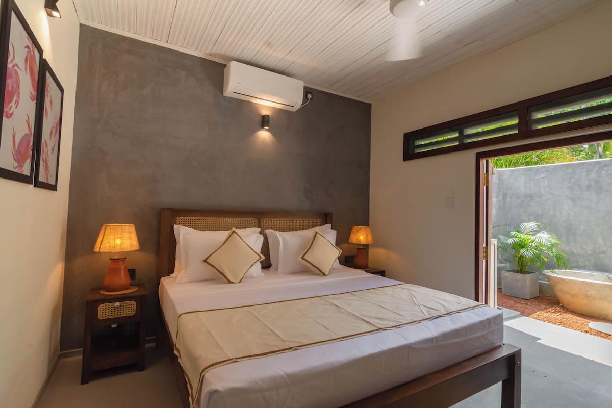 A bedroom with a bed, bedside lamps, a wall-mounted air conditioner, and a view of a garden through a sliding door.