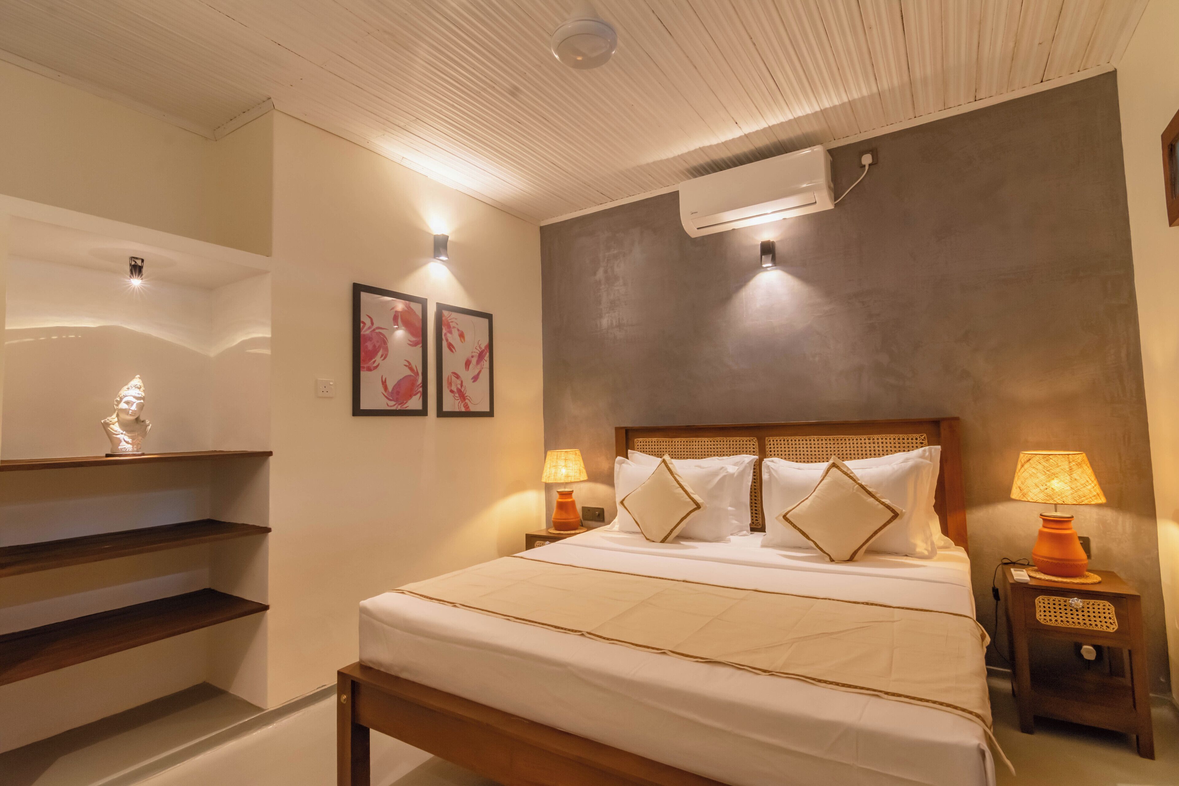 A neatly arranged bedroom with a bed, bedside lamps, a wall-mounted air conditioner, and a small shelf with a sculpture.