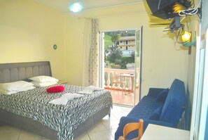 A bedroom with a bed, a sofa, a table, and a balcony with a view of buildings and greenery.