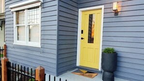 A house with a yellow door, a small porch with a potted plant, and a black metal fence. - "The Chadwick" Unique, High-end Boutique. (Soldiers Hill)