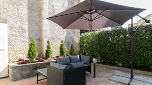 Um pátio externo com um sofá de vime preto, uma poltrona combinando e um grande guarda-sol. Há plantas em vasos e um hedge ao fundo.