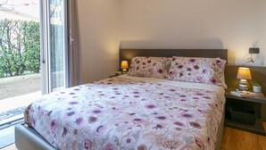 Quarto com cama, mesas de cabeceira, aparelho de ar condicionado e vista através de uma porta de vidro.