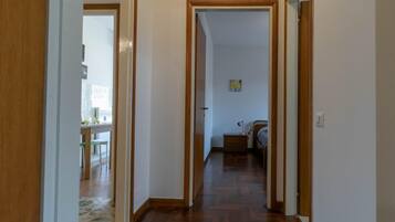 A hallway with wooden flooring and white walls, leading to a bedroom with a bed and a nightstand.