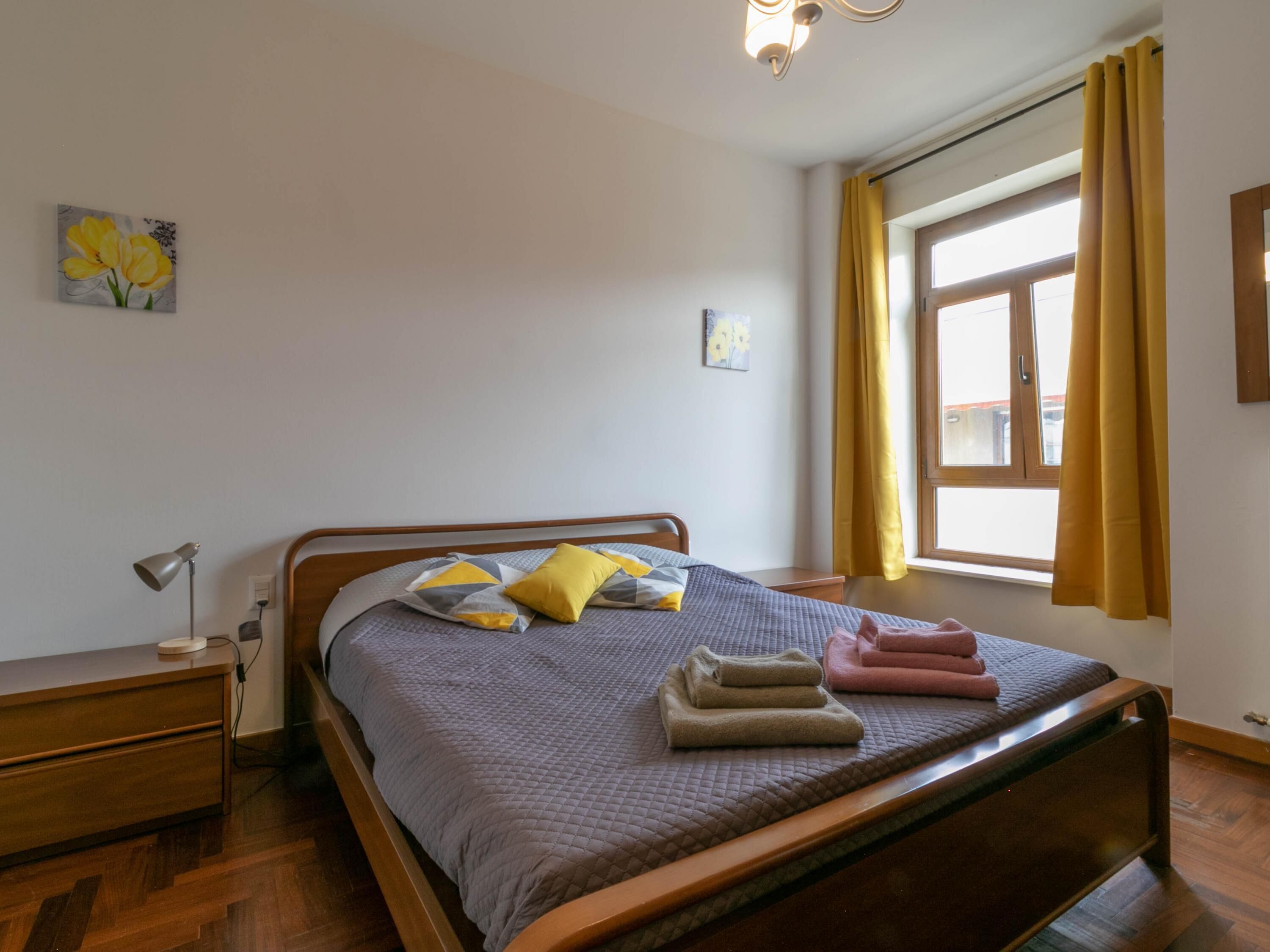 A neatly made bed with a gray quilt, two pillows, and folded towels. There is a bedside table with a lamp, a window with yellow curtains, and artwork on the wall.