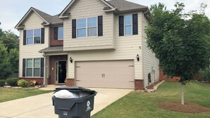 A two-story house with a garage, a front yard with a tree, and a black trash bin in the driveway.
