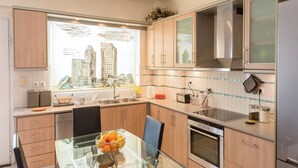 A modern kitchen with wooden cabinets, a glass dining table, and a window overlooking a cityscape. - Santa Barbara 2fl Home w. Garden and Intex Pool (Marathonas)