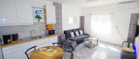 Una cocina moderna con armarios blancos, una zona de comedor con mesa y sillas de madera, y un sofá gris en un espacio habitable luminoso con piso de baldosas.