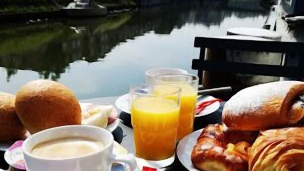 Pöydällä on aamiaistarvikkeita, kuten croissantteja, kuppi kahvia ja appelsiinimehua, ja taustalla näkyy kanava sekä sen varrella olevat rakennukset.