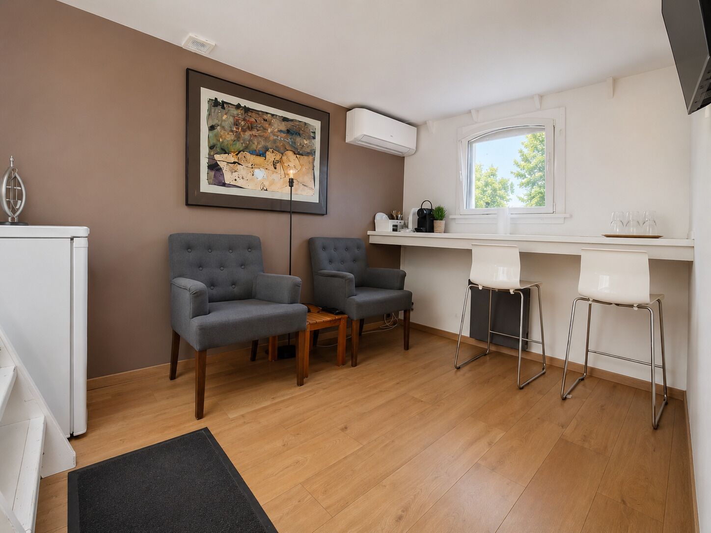 A modern living room with two grey armchairs, a wooden coffee table, a black rug, a white desk with a computer, and a window with a view of trees.
