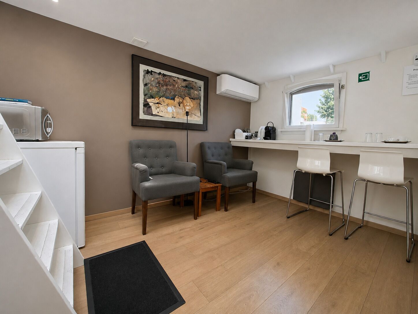 Een moderne kamer met een kitchenette, twee grijze fauteuils, een houten salontafel, een zwart vloerkleed en een schilderij aan de muur.