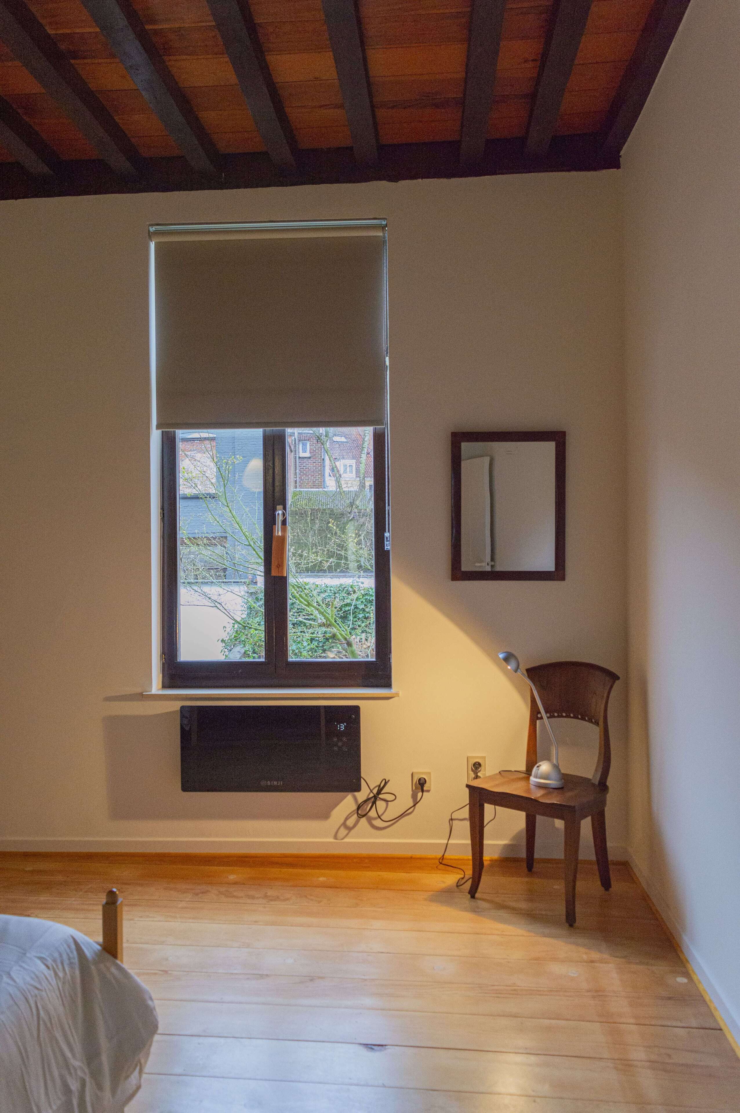 Une pièce avec un plafond en bois, une fenêtre avec un store, une chaise, une lampe de bureau et un miroir fixé au mur.
