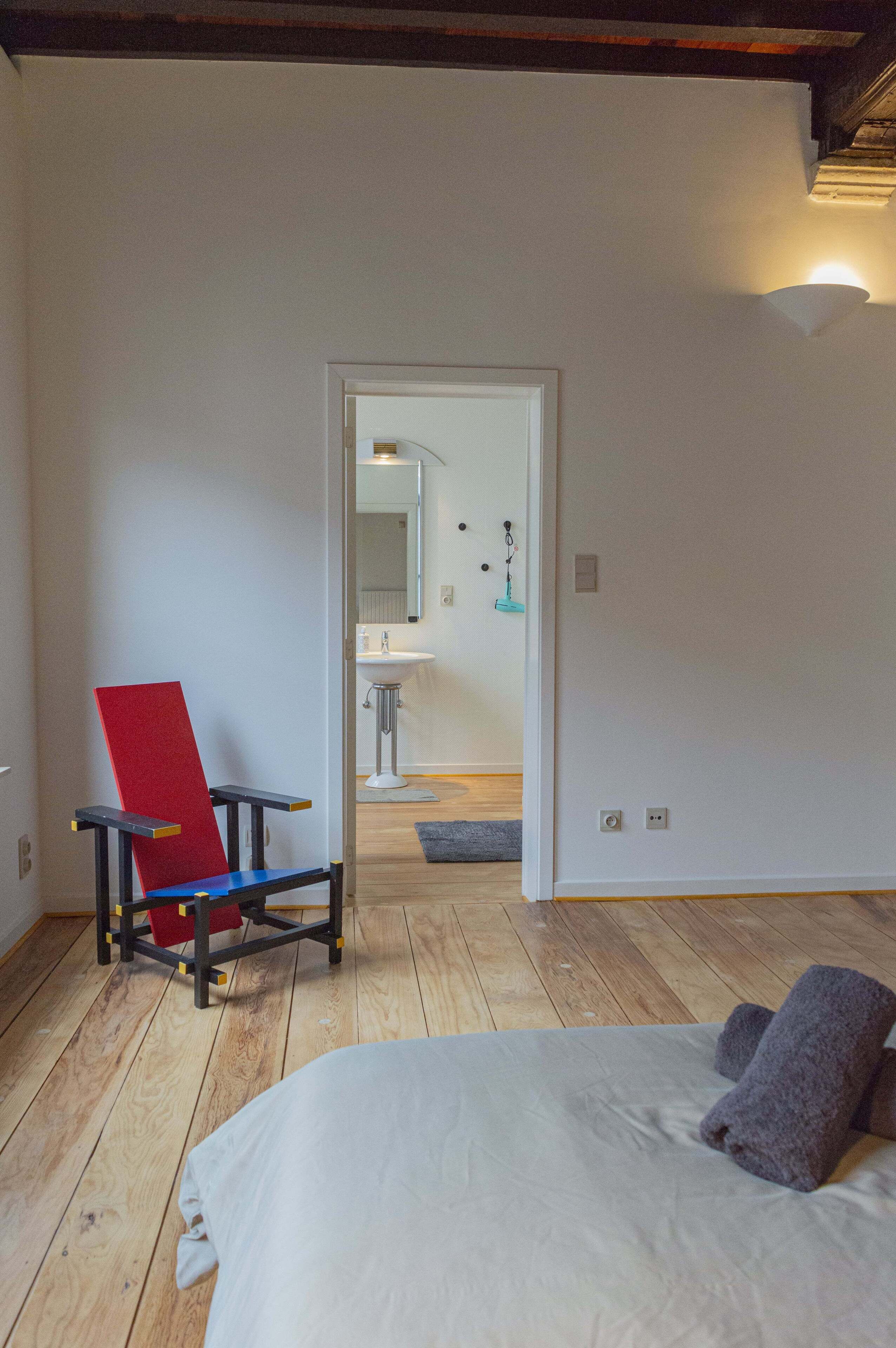Una camera da letto con un letto, una sedia rossa e blu e vista sul bagno.