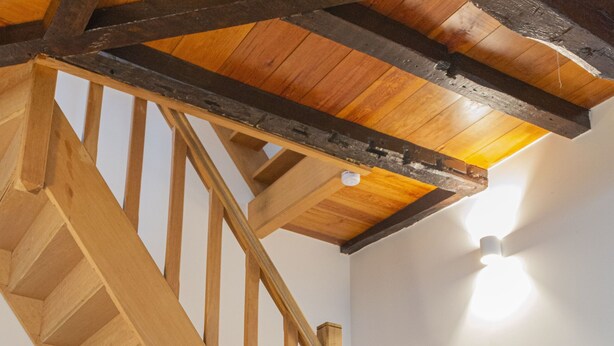 Eine Holzt Treppe mit Handlauf, Holzbalken an der Decke und einer Wandleuchte.