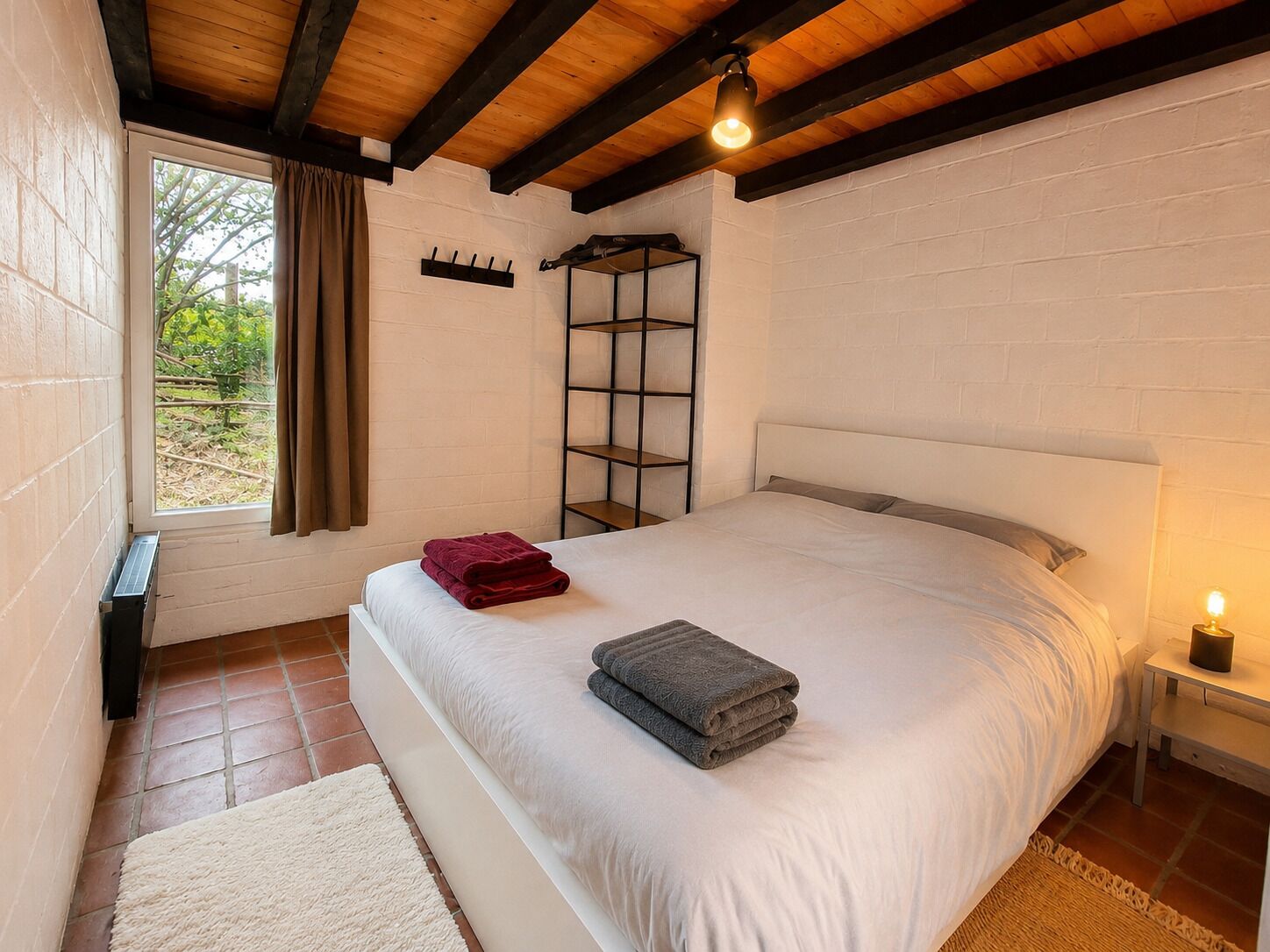 A bedroom with a bed, a nightstand, a window with curtains, and a shelf unit.