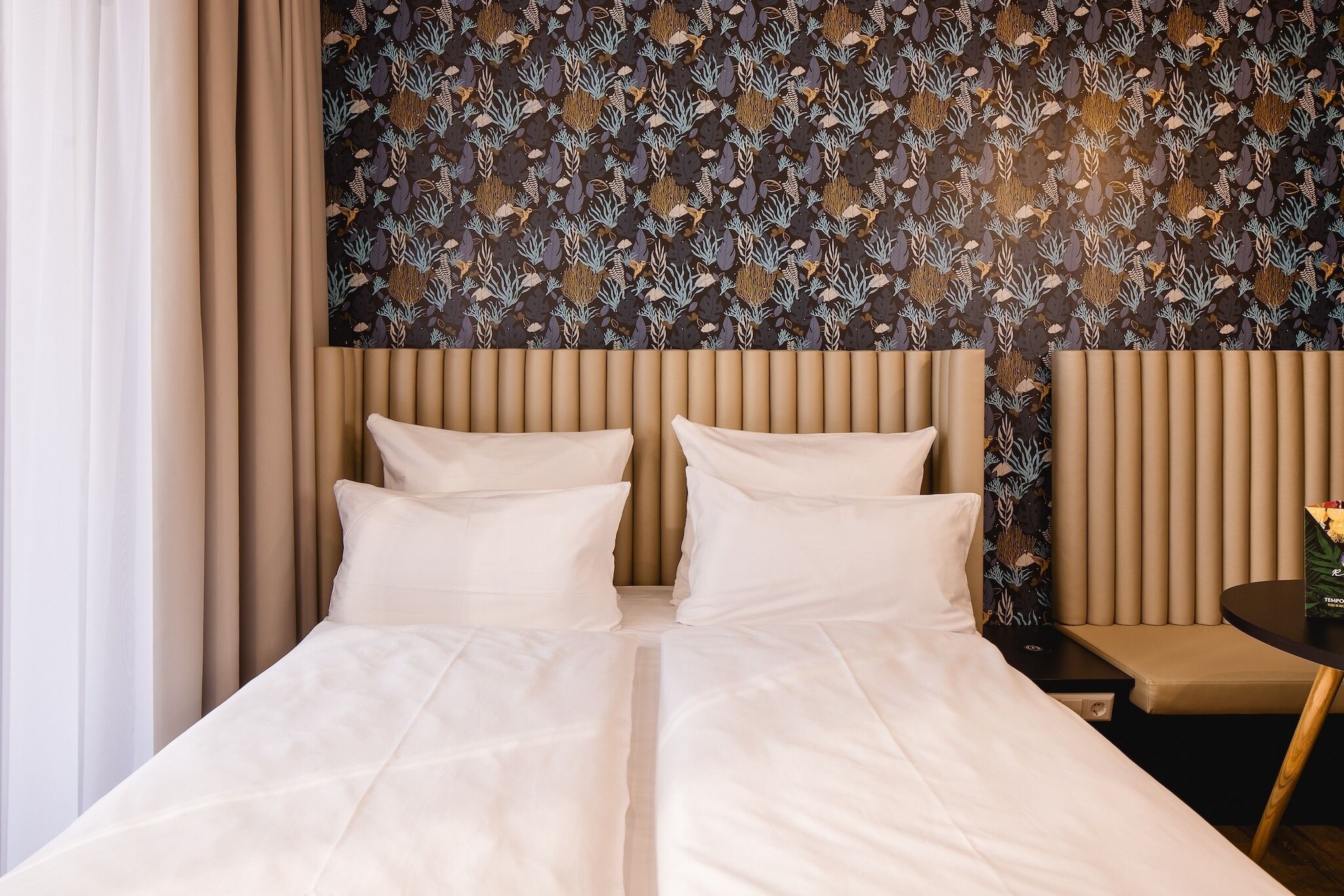 A hotel room with a large bed, two pillows, a headboard with vertical slats, a patterned wallpaper, and a small round table with a vase.