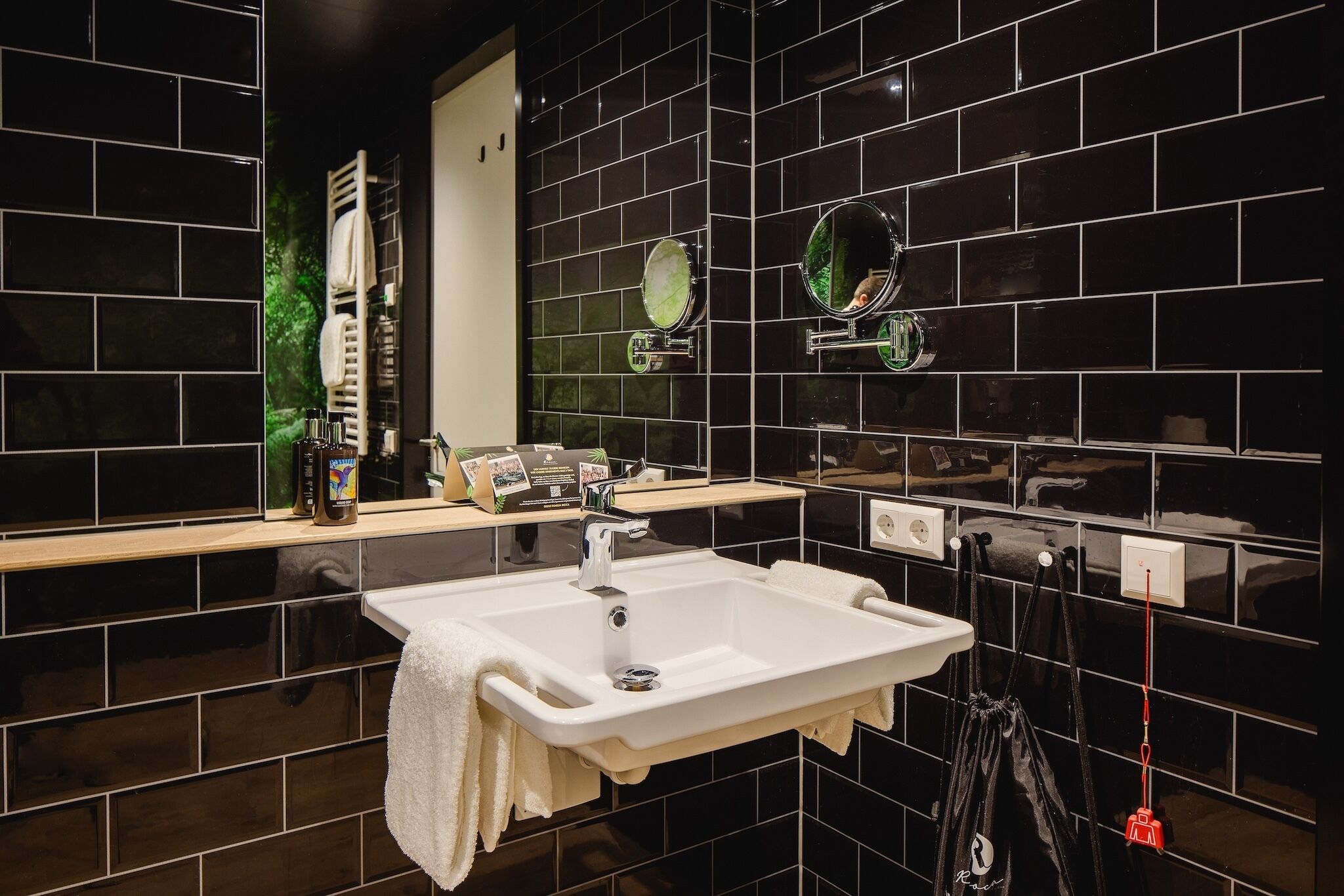 Une salle de bain moderne avec des murs recouverts de carreaux noirs, un lavabo blanc et un miroir agrémenté de deux petits miroirs ronds.