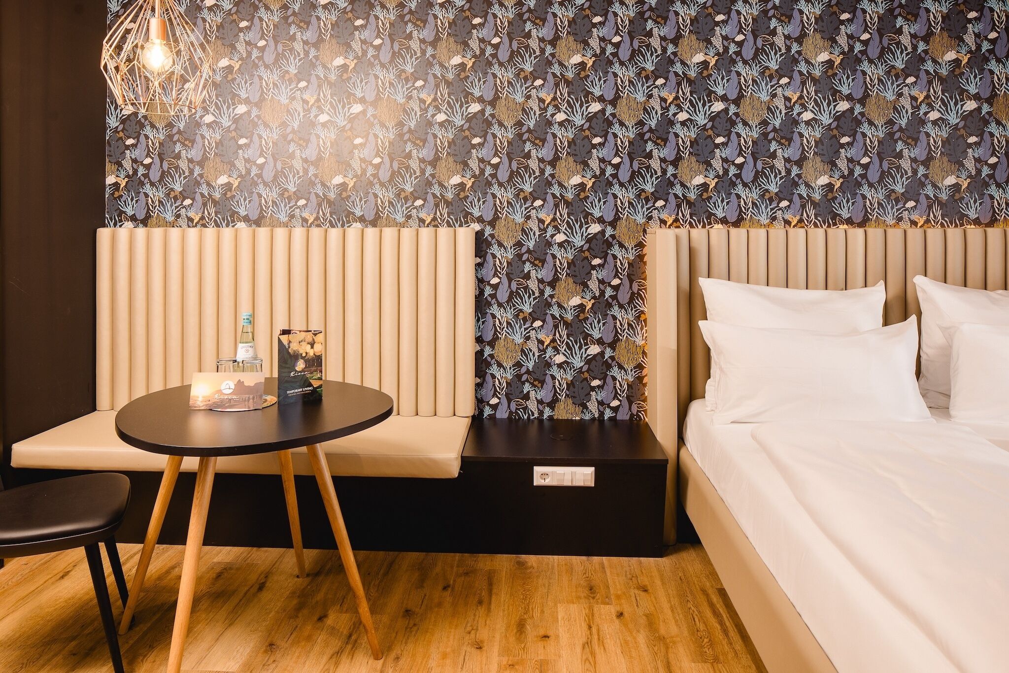 A modern hotel room with a bed, a round table with a chair, a small sofa, and a patterned wallpaper.