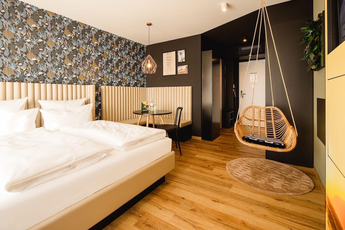 A modern bedroom with a large bed, a hanging chair, a small table, and a patterned wall.