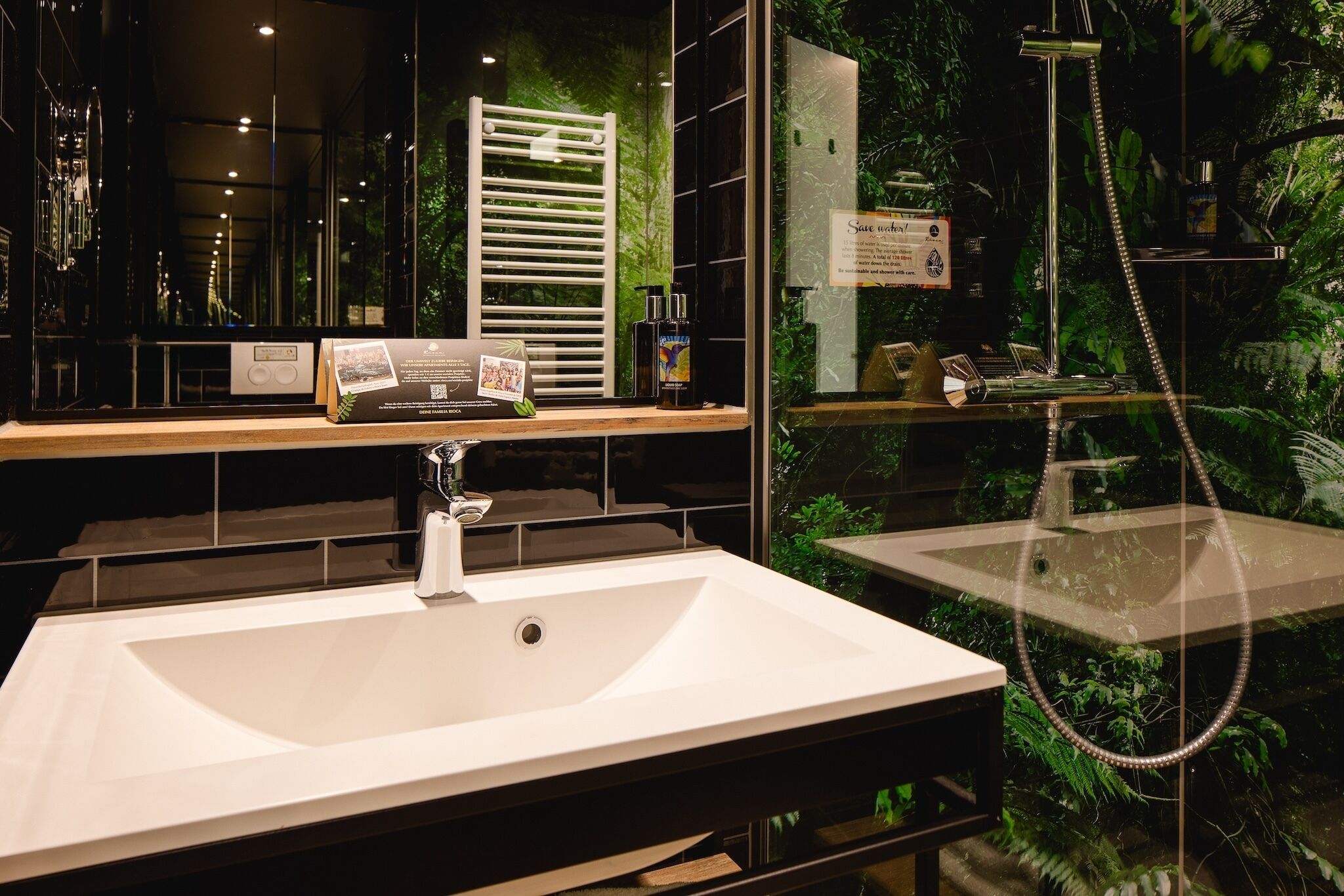 Un baño moderno con un lavabo grande de color blanco, un cabezal de ducha manual y una zona de ducha con mampara de cristal y plantas.