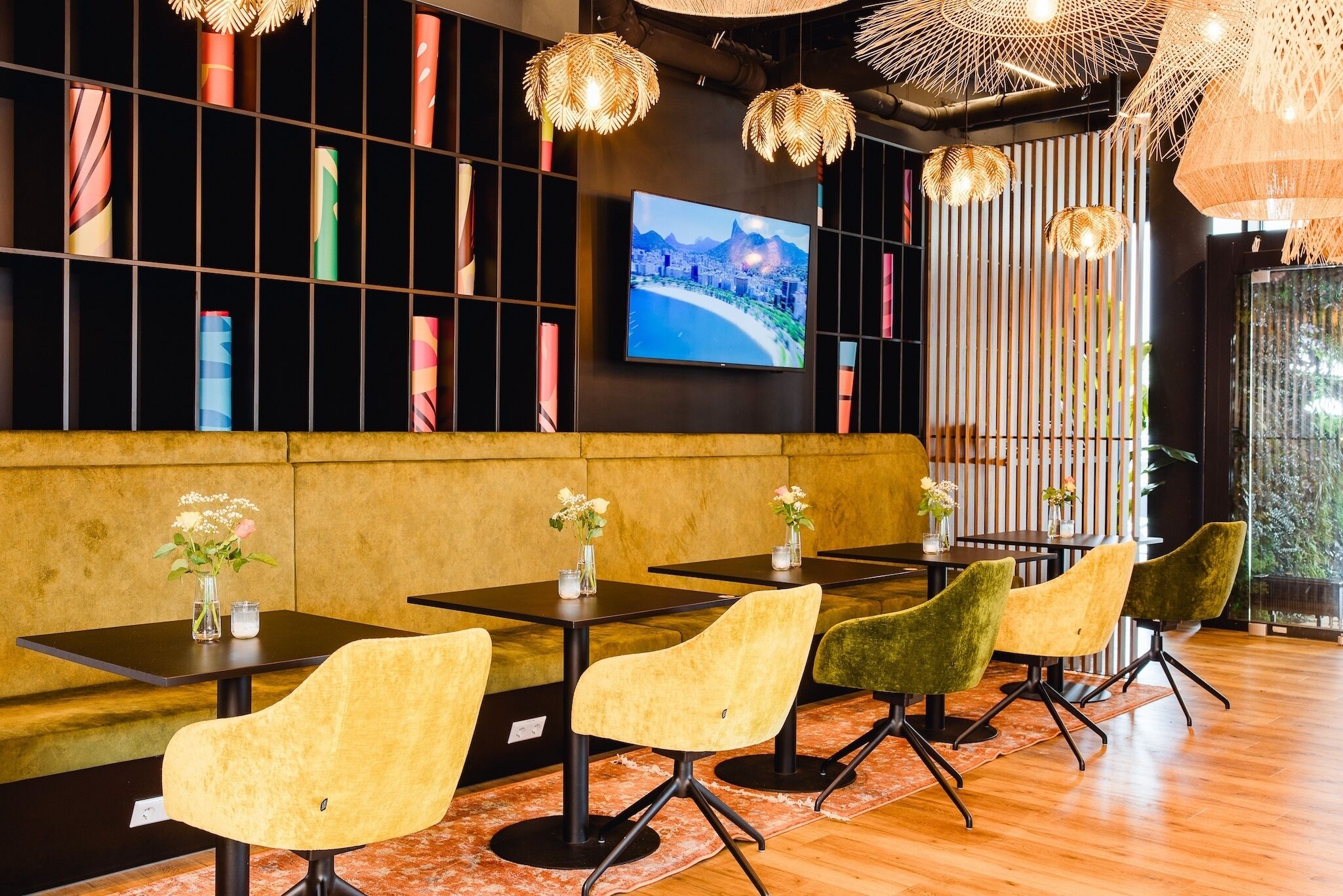 A modern restaurant with black tables, yellow and green chairs, and a TV displaying a scenic view.