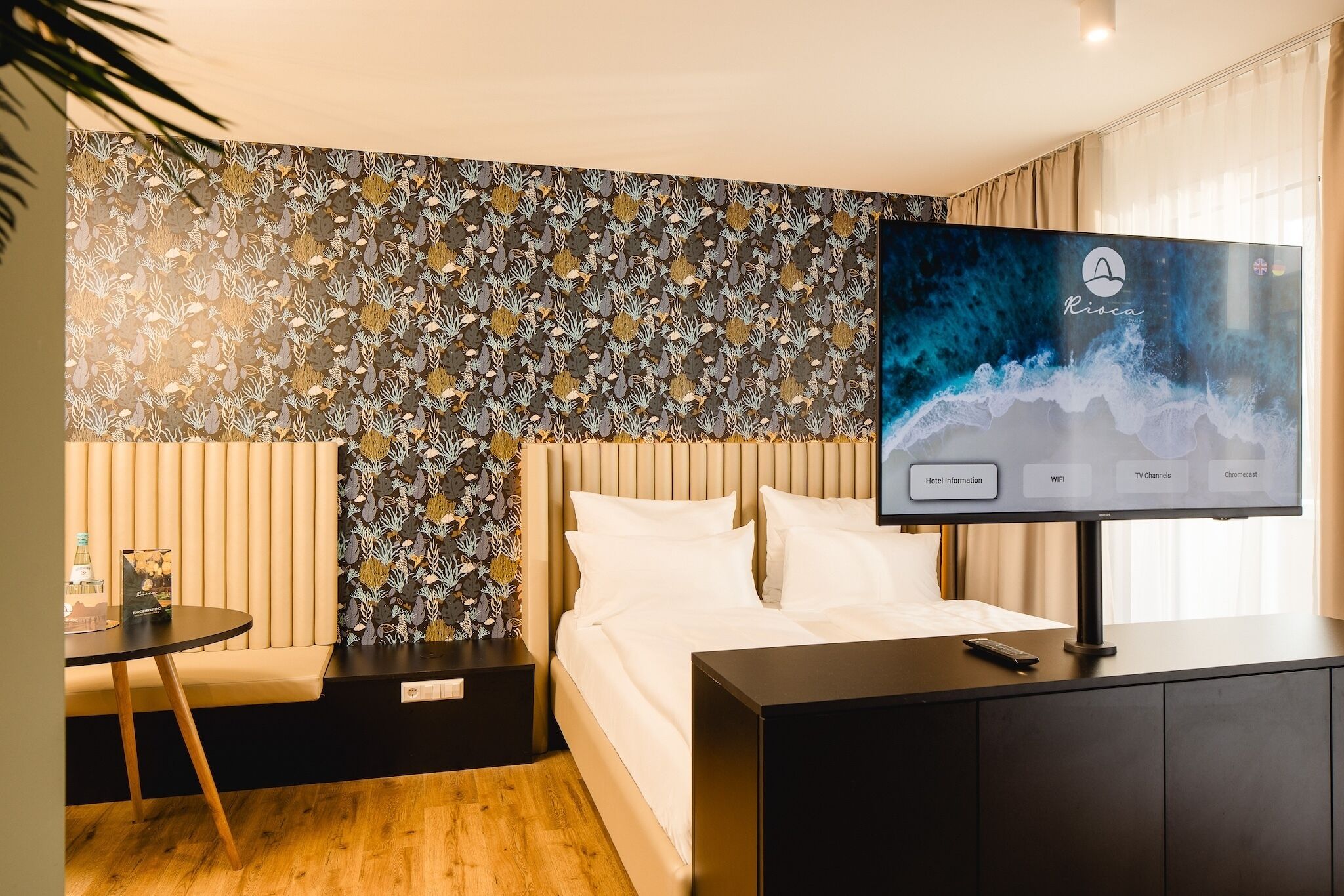 A hotel room with a bed, a TV on a stand, a small table, and a decorative wall.