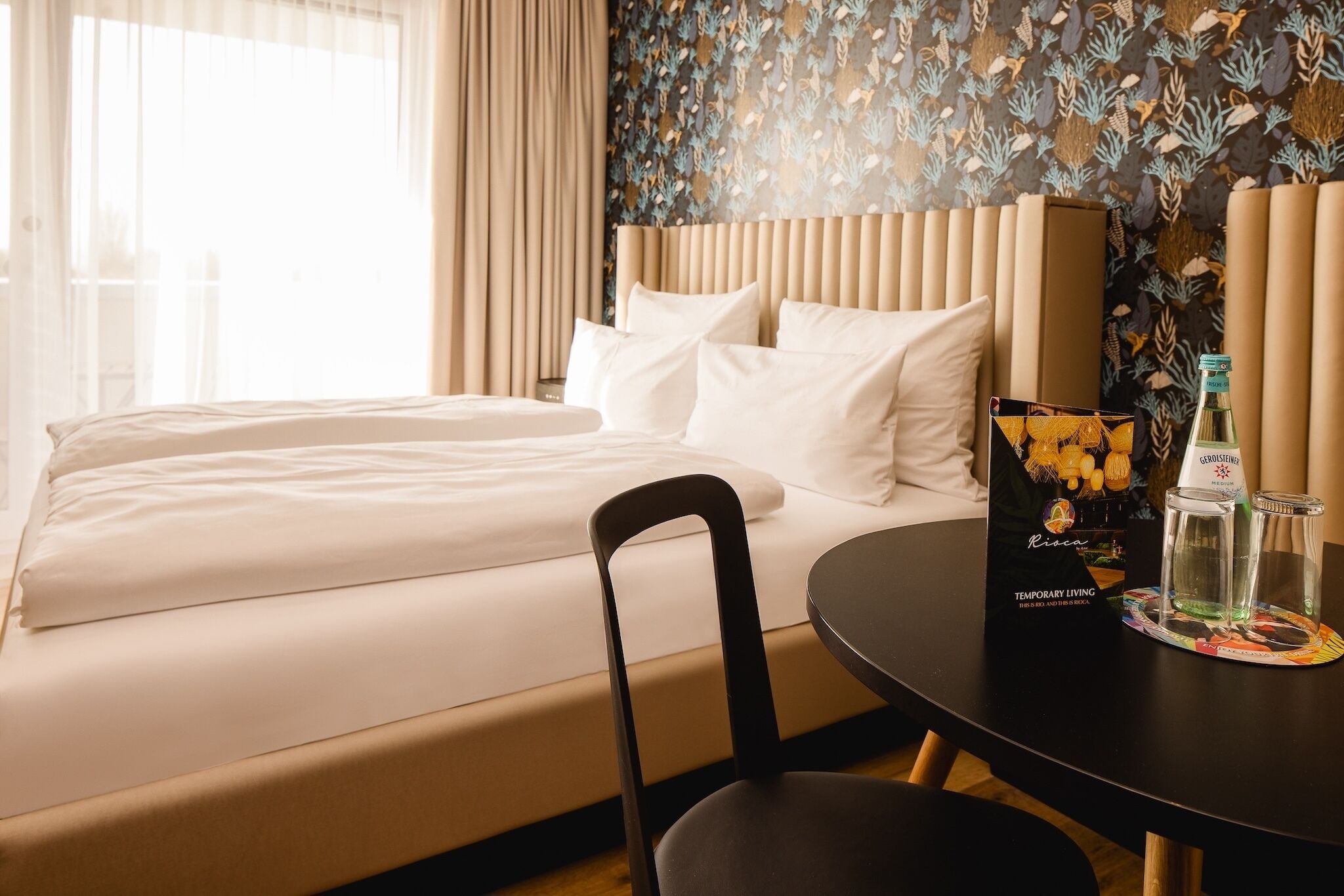 Une chambre d’hôtel avec un lit, une chaise, une table sur laquelle se trouvent une bouteille et un verre, ainsi qu’un objet décoratif.