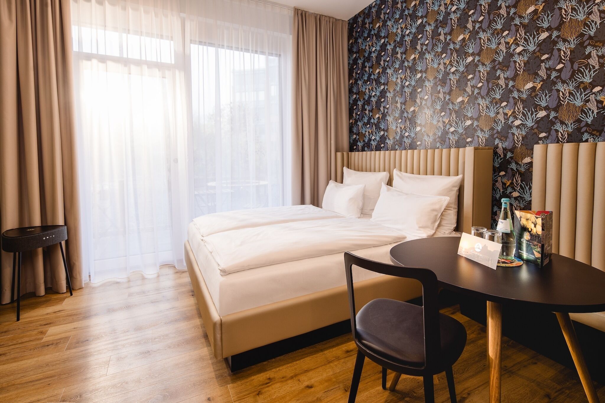Hotellihuone, jossa on sänky, pyöreä pöytä ja tuoli, pieni sivupöytä ja kuviollinen seinä.